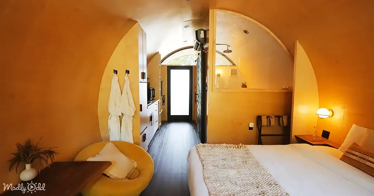 A cozy, warmly lit bedroom with a bed, yellow chair, robe hooks, and a small bathroom with a shower.