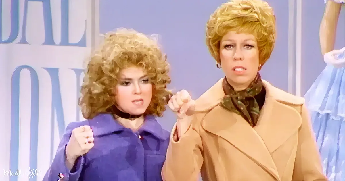 Two women with large, curly blonde hair, one in a tan coat and the other in a blue coat, appear to be on a talk show set.