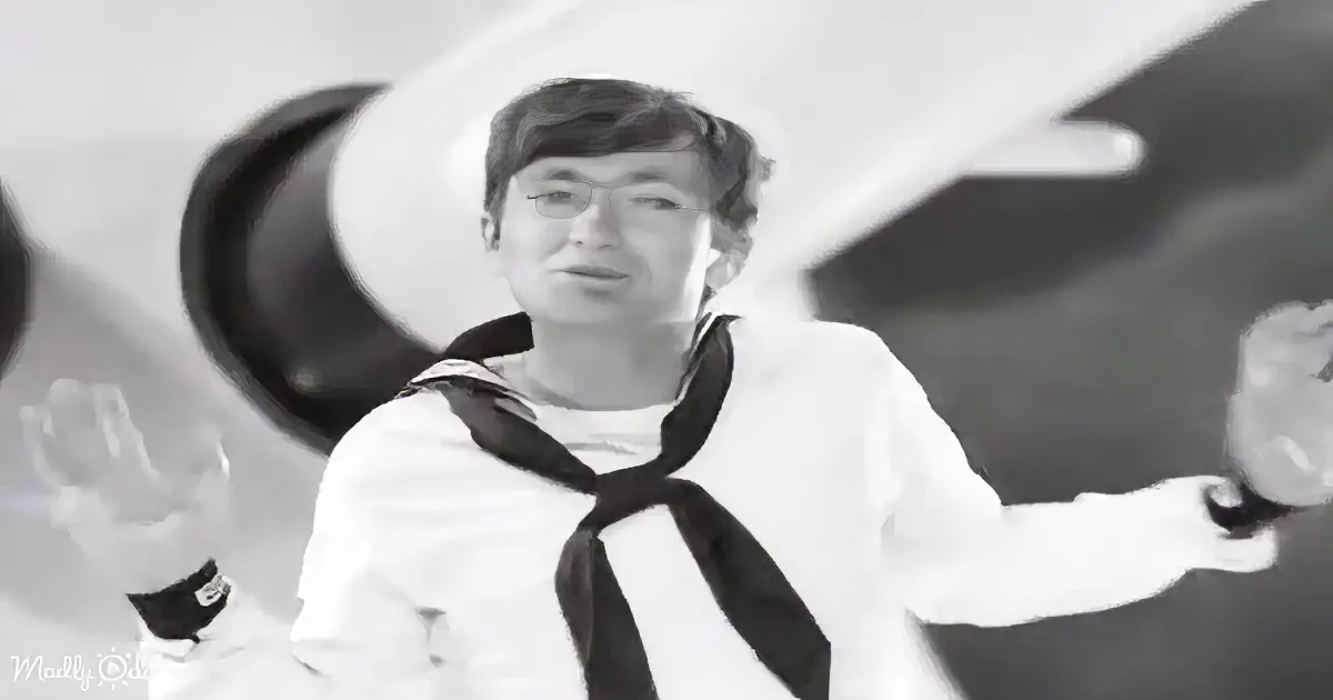 A person in a sailor uniform is waving and holding a small object, with a blurred background.