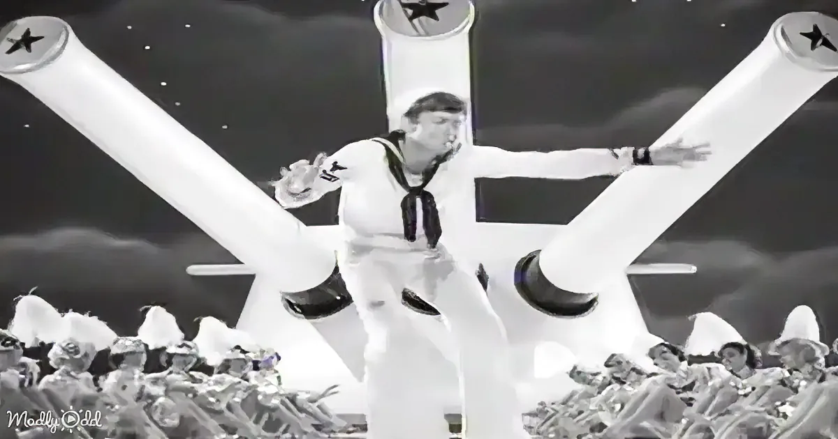 A sailor in a white uniform stands with arms outstretched, surrounded by a crowd of uniformed figures, with large...