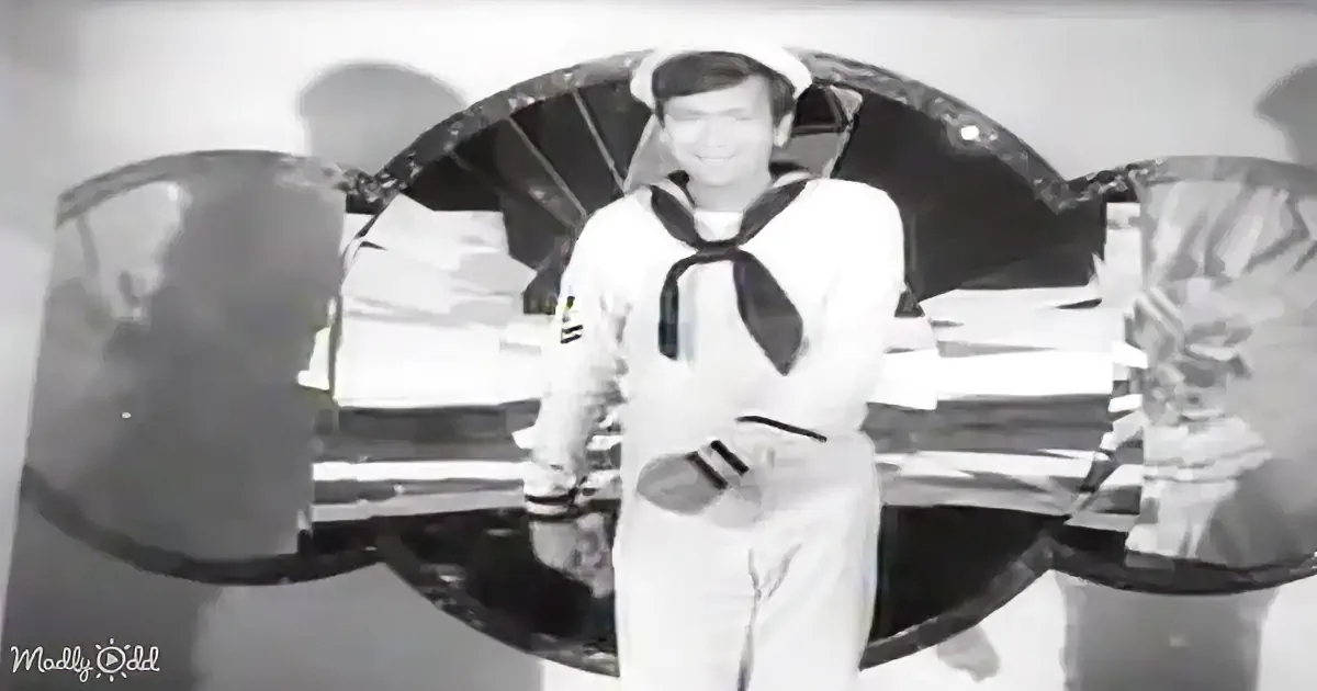 A sailor in a white uniform stands on a ship, surrounded by large, cylindrical objects.