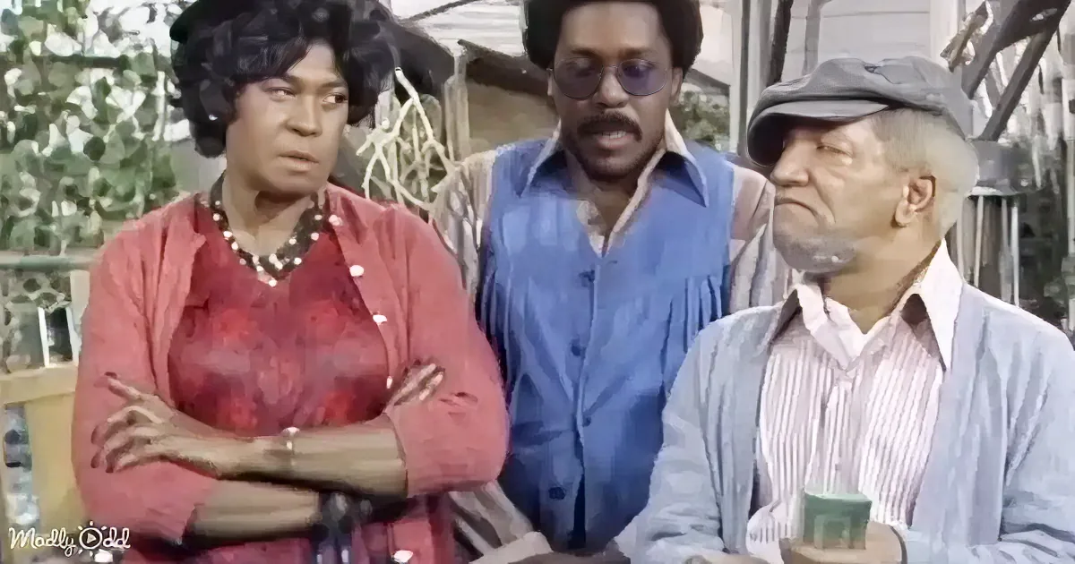 A woman in a red dress with her arms crossed stands next to a man in a blue shirt and another man in a striped shirt.
