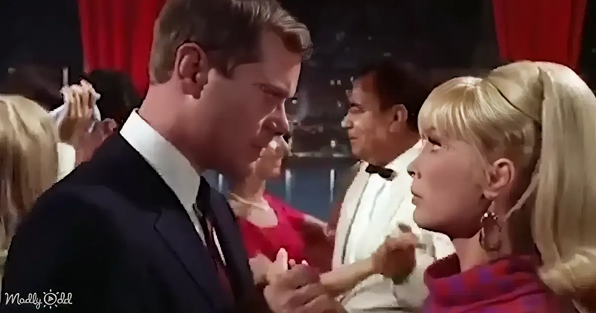 A man in a suit is holding hands with a woman in a red dress, surrounded by people in a social setting.