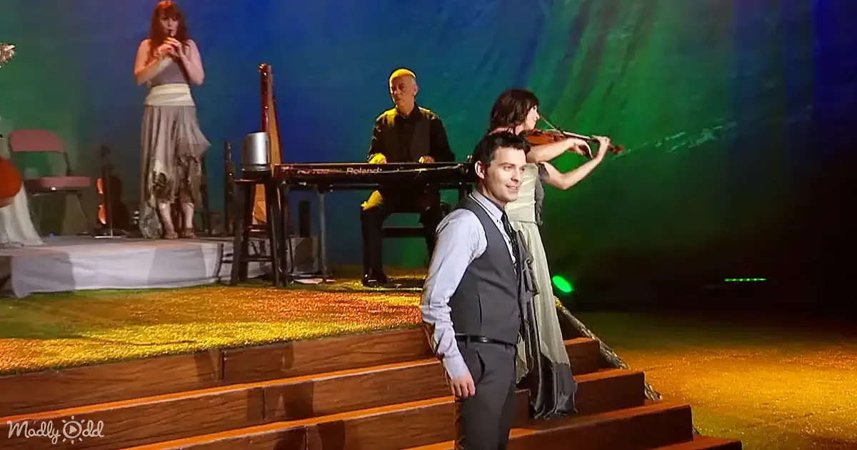 A man in a suit stands on stage with musicians playing instruments behind him.