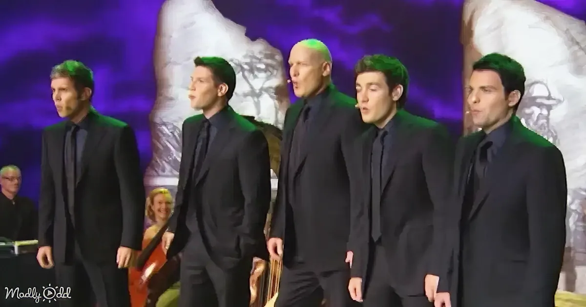 Five men in black suits sing on stage with a purple backdrop and musicians in the background.