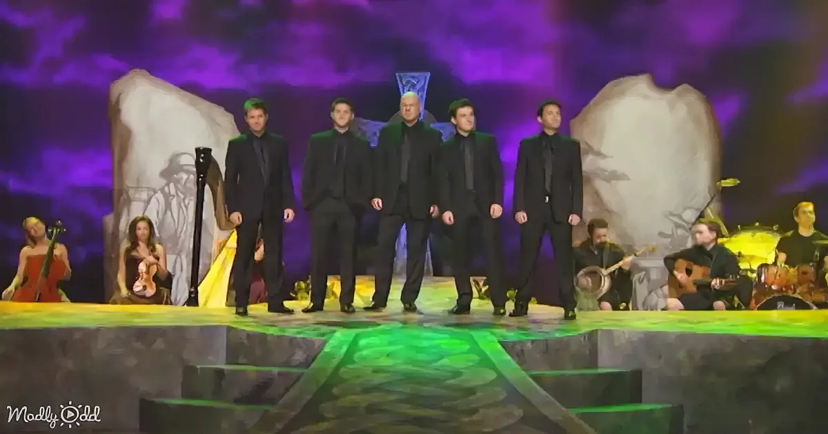A group of men in black suits stands on stage with musicians and a harpist behind them.