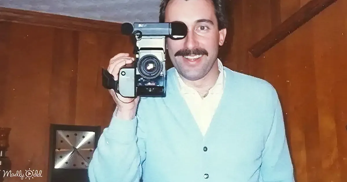 A man in a light blue cardigan holds a vintage camera to his eye, smiling.