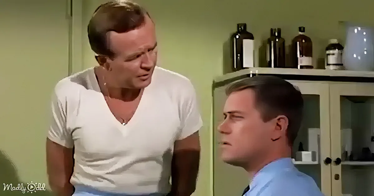A man in a white shirt leans over another man in a blue shirt, seemingly in a medical or professional setting.