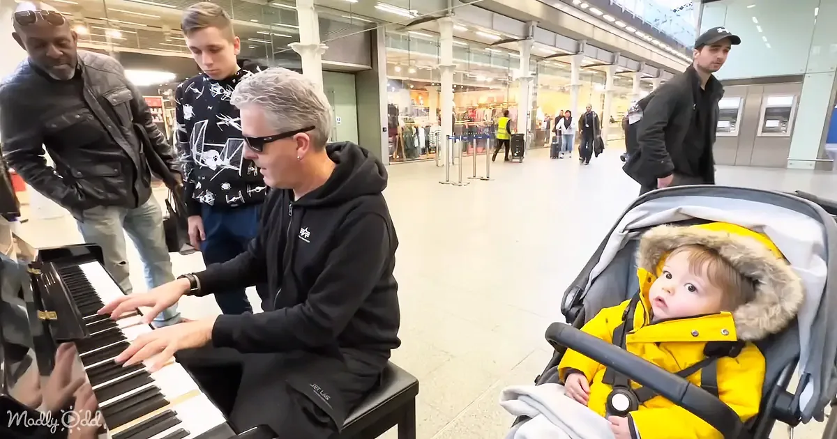 A man in a black hoodie plays a piano in a busy airport, surrounded by onlookers, with a baby in a yellow coat sitting in...