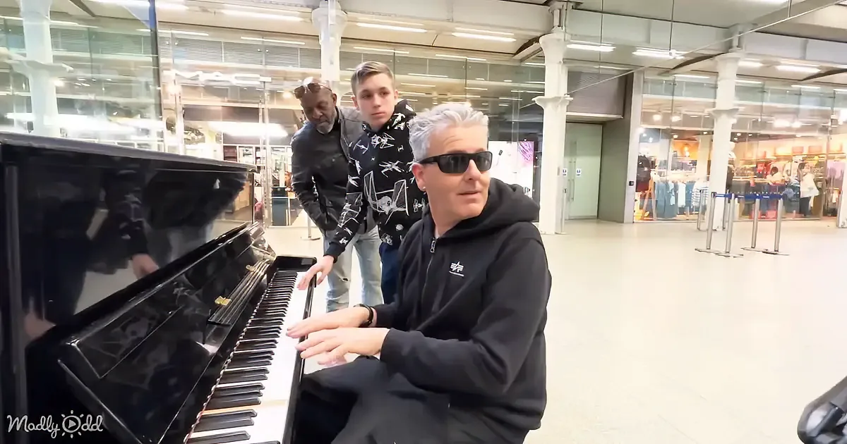 A man in sunglasses plays a piano in a spacious indoor area with two others observing.