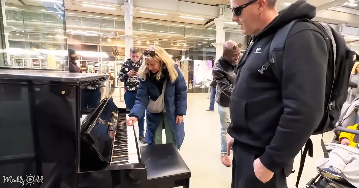 A woman in a blue coat plays a black piano in a busy indoor setting with onlookers.