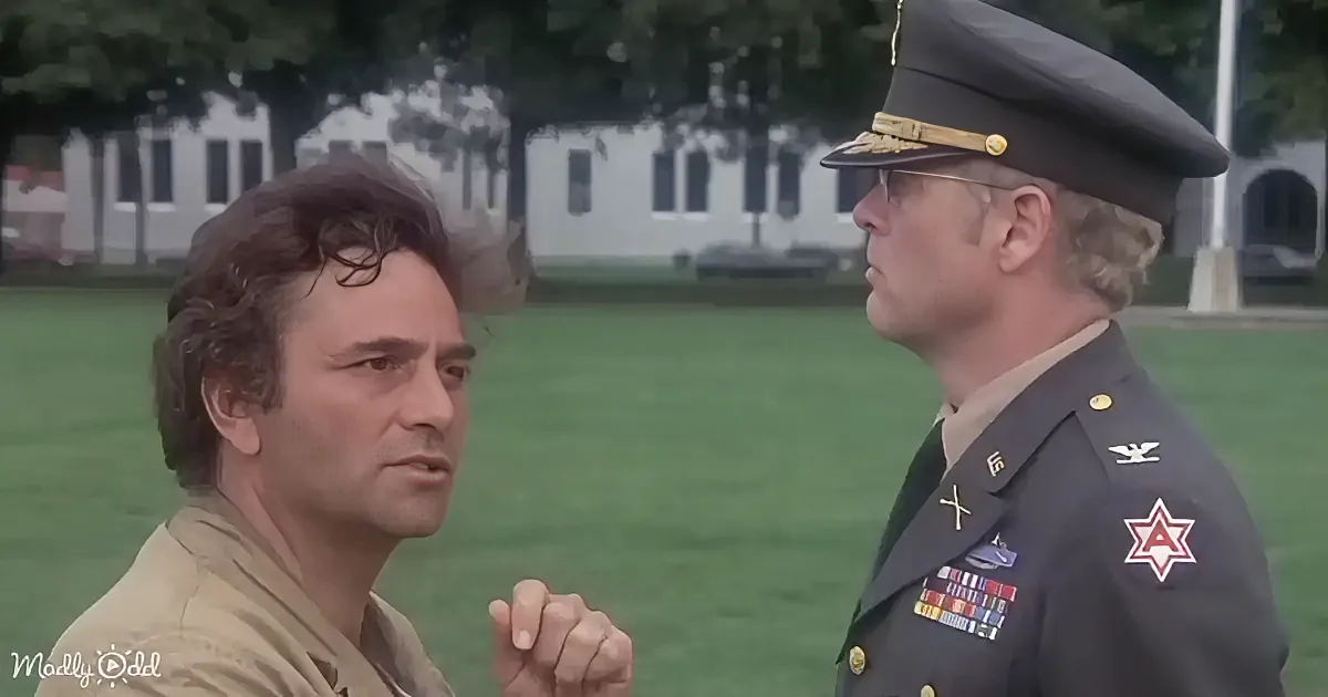 A man in a military uniform speaks to another man in a tan shirt, both outdoors with a building and trees in the background.