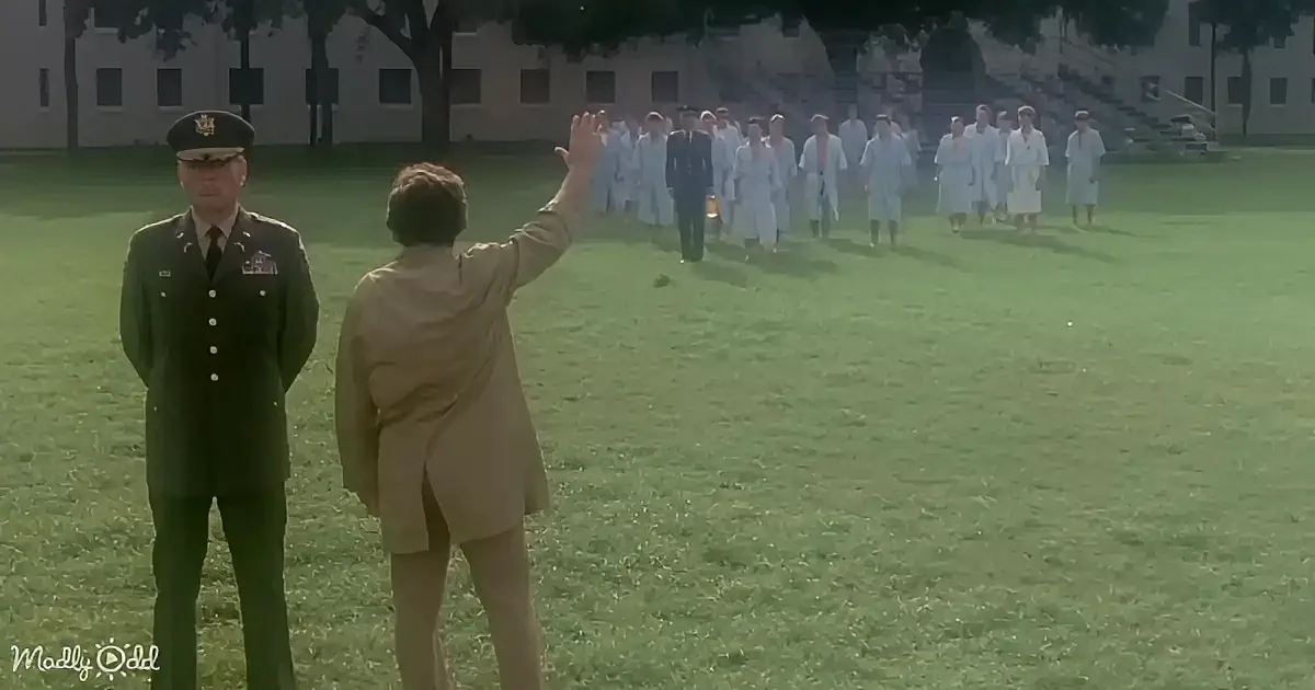 A woman in a beige suit waves to a group of people in white coats walking on a grassy field.