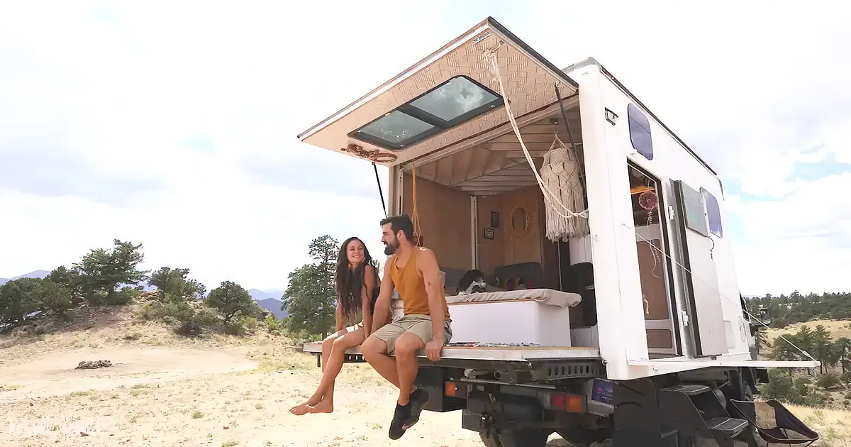 A couple sits on the back of a white camper truck in a desert landscape, enjoying the view.
