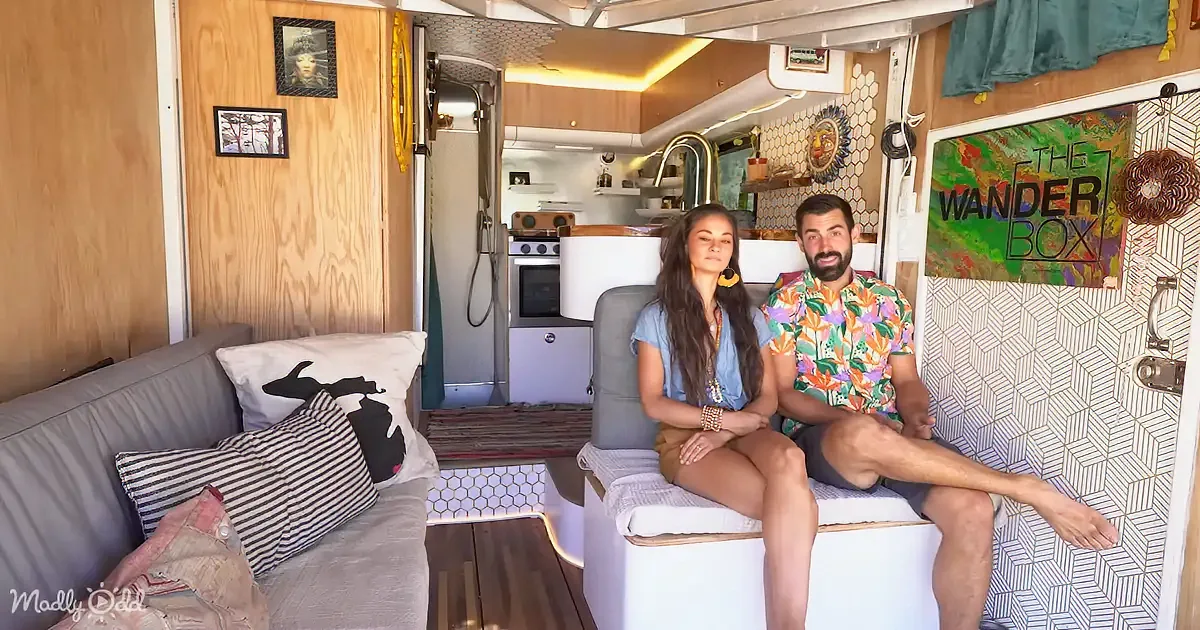 A couple sits on a bed in a cozy, well-decorated RV with a kitchen and hexagonal tiles.