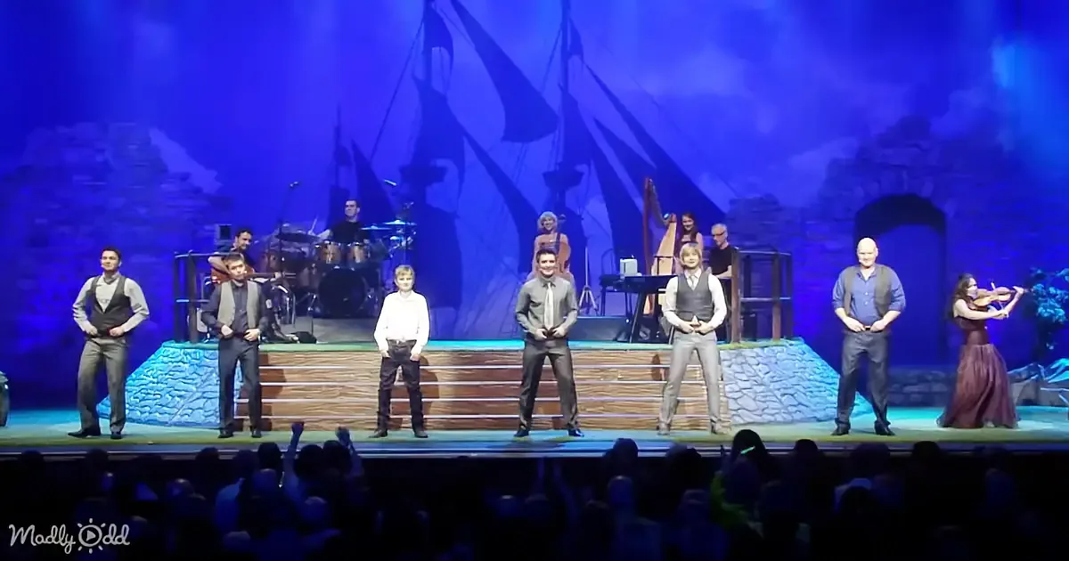 A group of men in formal attire stands on a stage with musicians and a harpist in the background.