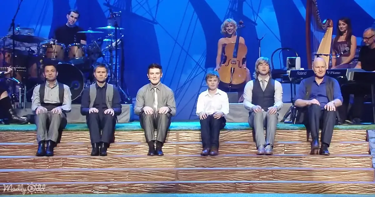 Six men in formal attire sit on stage steps, with musicians and instruments behind them.