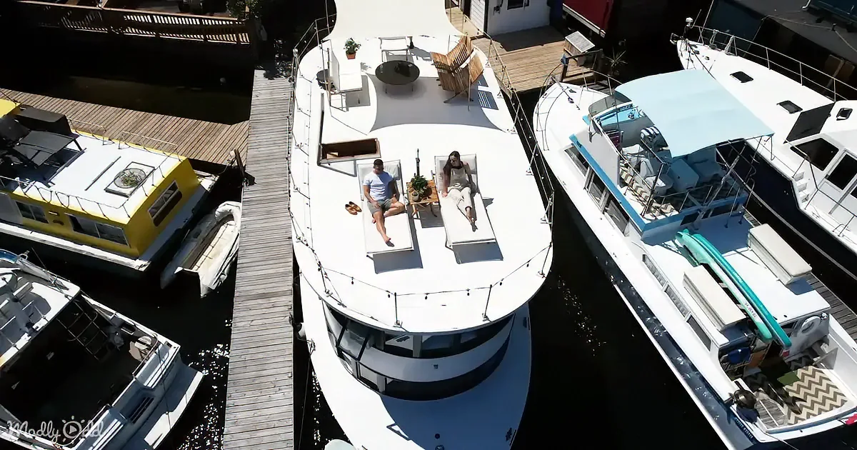 A couple lounges on a white houseboat's deck surrounded by other boats and a dock.