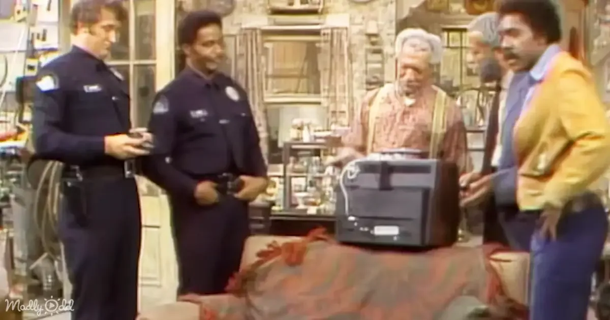 Police officers and a man in a yellow jacket stand in a room with a TV and furniture.