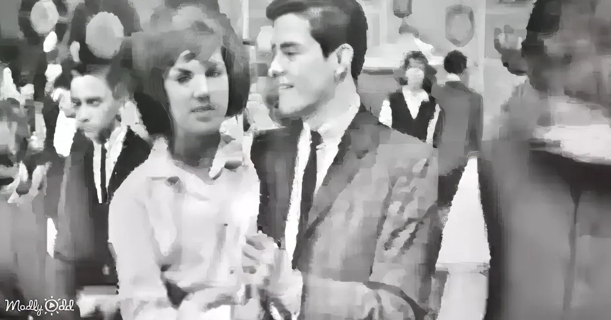 A man in a suit and tie stands next to a woman in a white blouse in a crowded room.