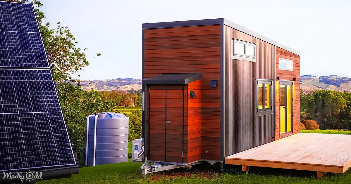 A modern tiny house with solar panels, a water tank, and a wooden deck sits on a grassy hillside.