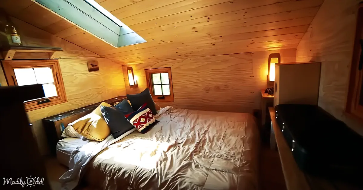 A cozy bedroom with wooden walls, a bed with pillows, a skylight, and a small window letting in natural light.