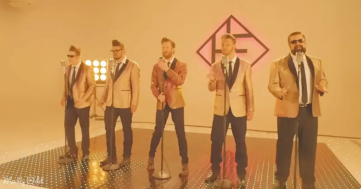 Five men in suits sing on stage with microphones, wearing sunglasses, under warm lighting.