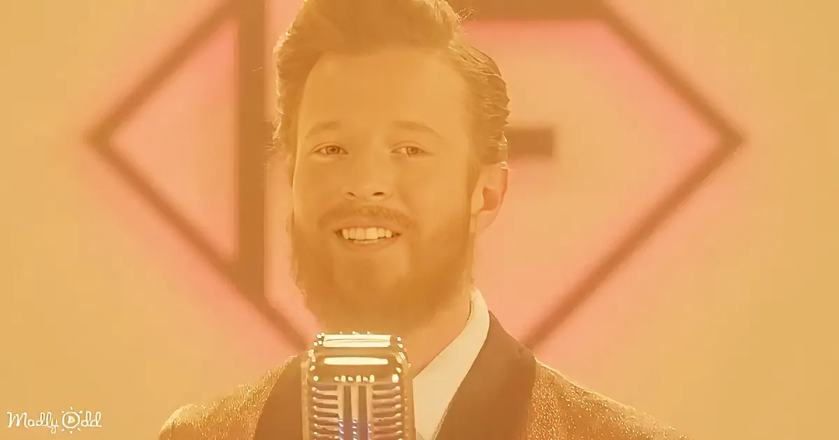 A man with a beard and slicked-back hair smiles while singing into a vintage microphone.