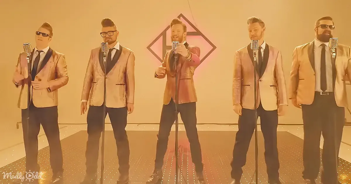 A man in a pink suit sings into a microphone, surrounded by four other men in similar attire, under warm lighting.