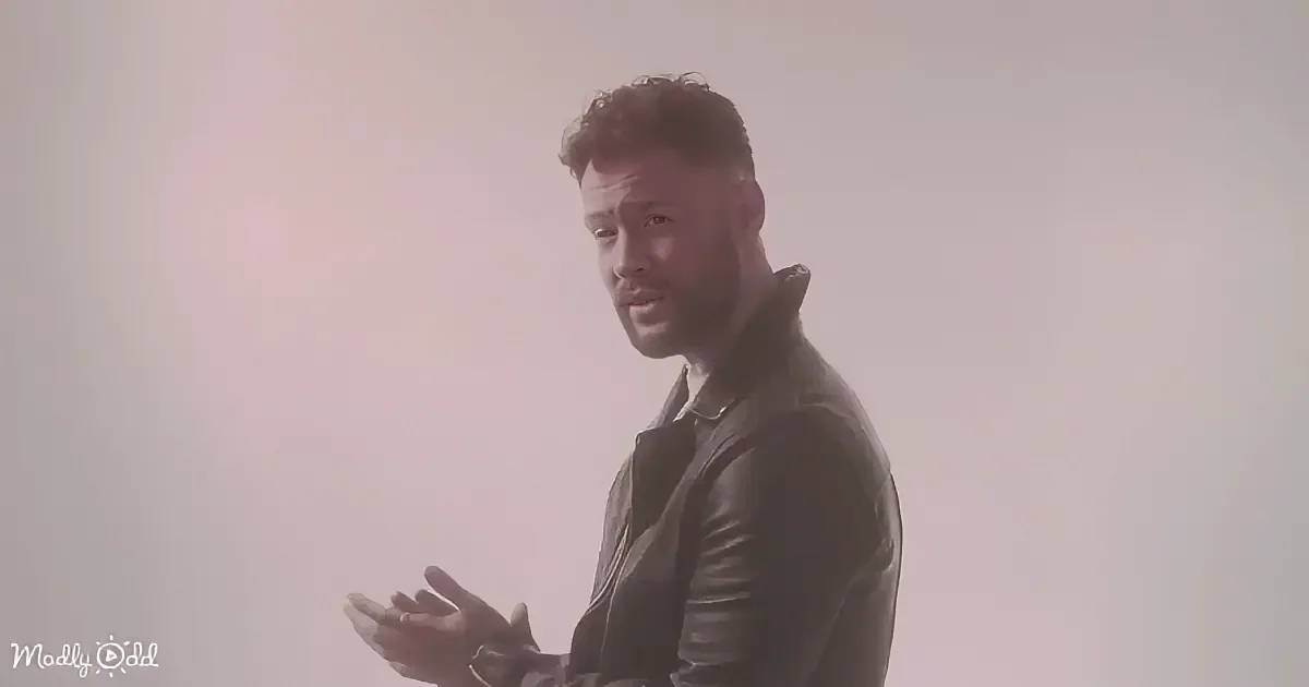 A man in a black leather jacket stands with hands clasped, looking to the side against a plain background.