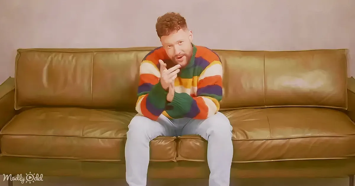A man in a colorful striped sweater sits on a brown leather couch, gesturing with his hands.