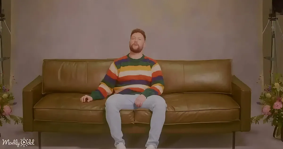 A man in a colorful striped sweater sits on a brown leather couch, surrounded by flowers.