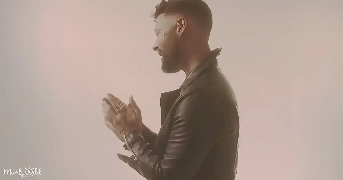 A man in a leather jacket stands with hands clasped, looking down, against a soft, blurred background.