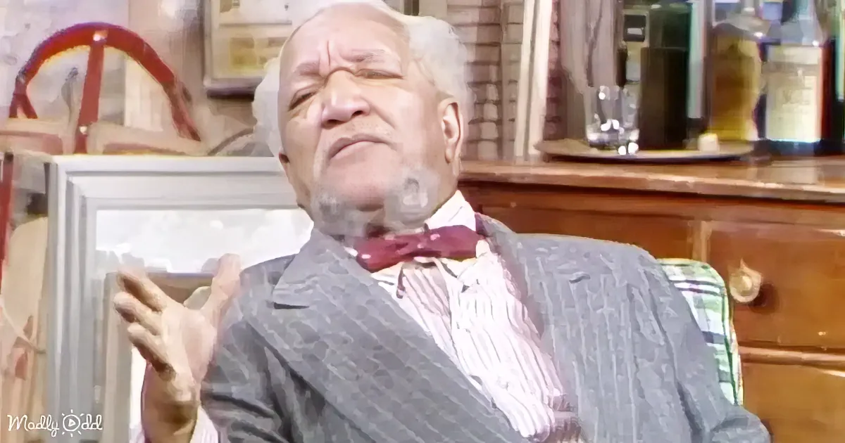 An older man in a suit and bow tie is sitting, gesturing with his hand, in a room with a wooden dresser and bottles.