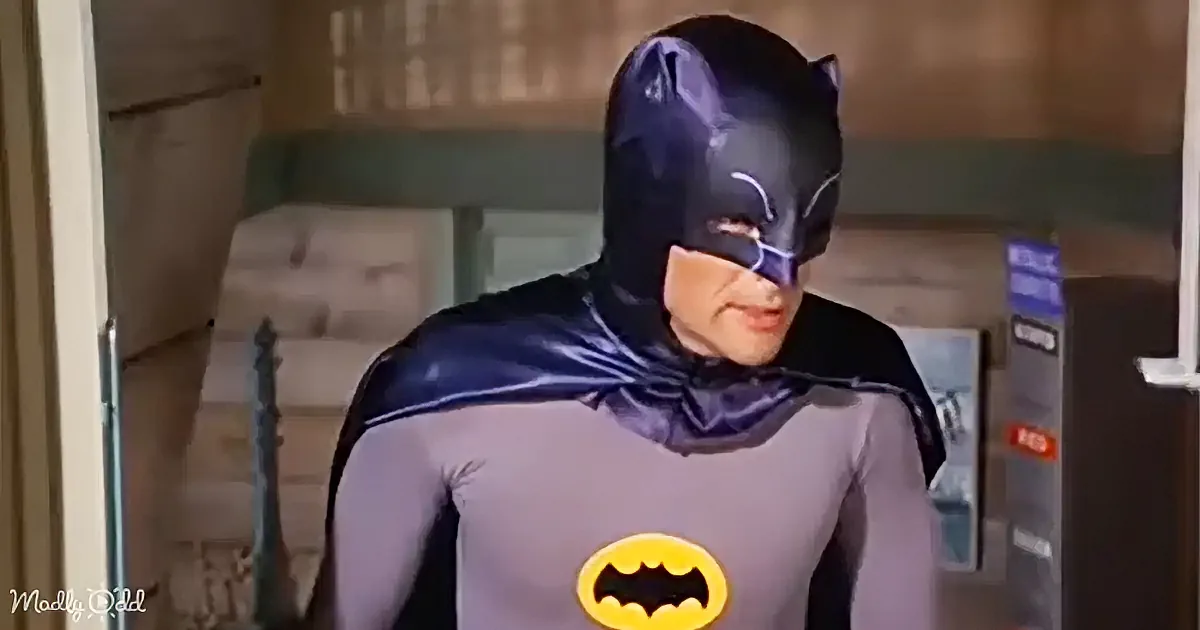 A person in a Batman costume stands in front of a door, wearing a gray suit with a yellow bat symbol.