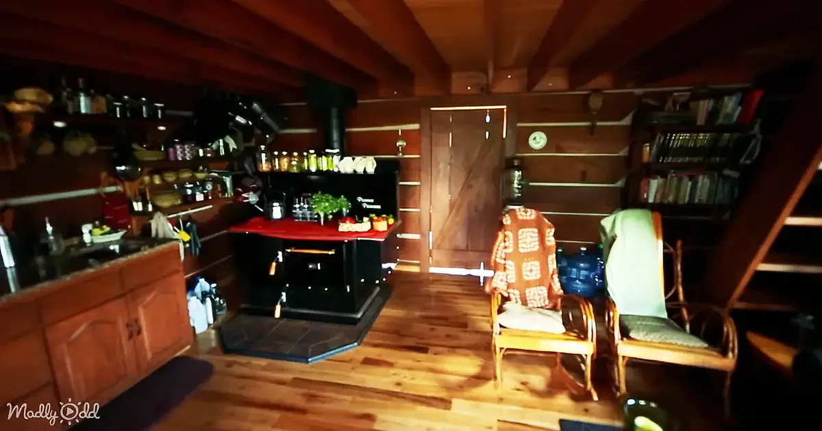 A cozy cabin interior with wooden floors, shelves, a stove, and two chairs.
