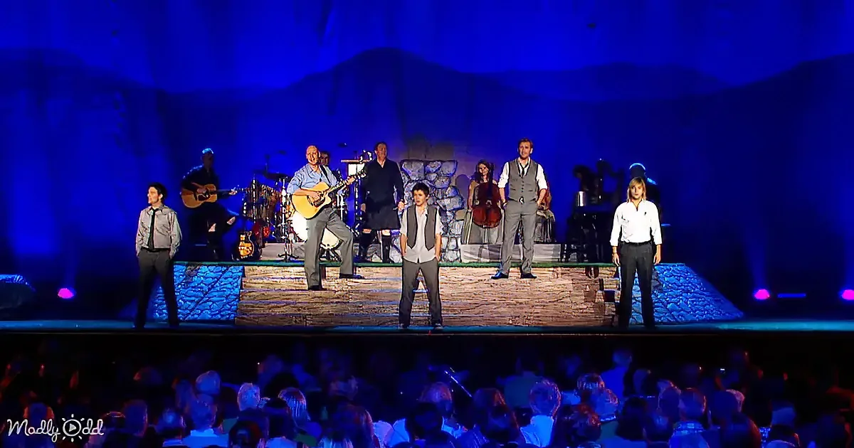 A group of performers and musicians on stage with a blue backdrop, illuminated by stage lights.