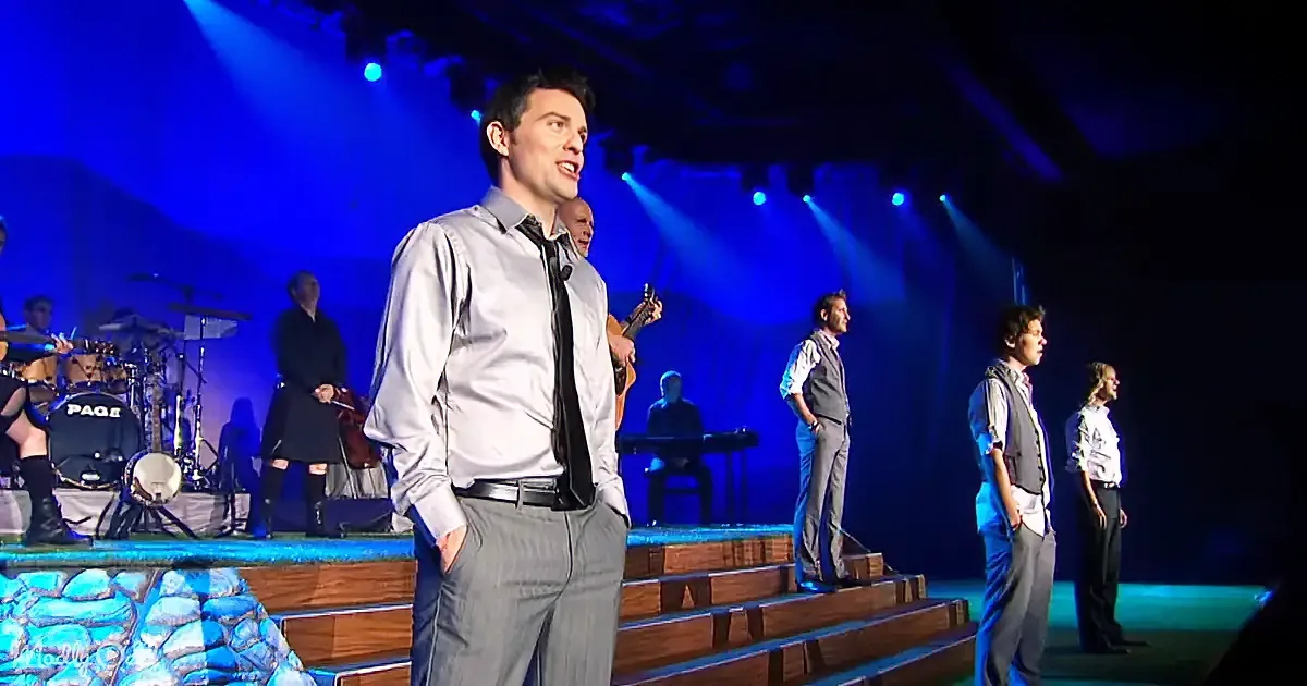 A man in a gray shirt and tie stands on stage, singing, with a band and other performers behind him.