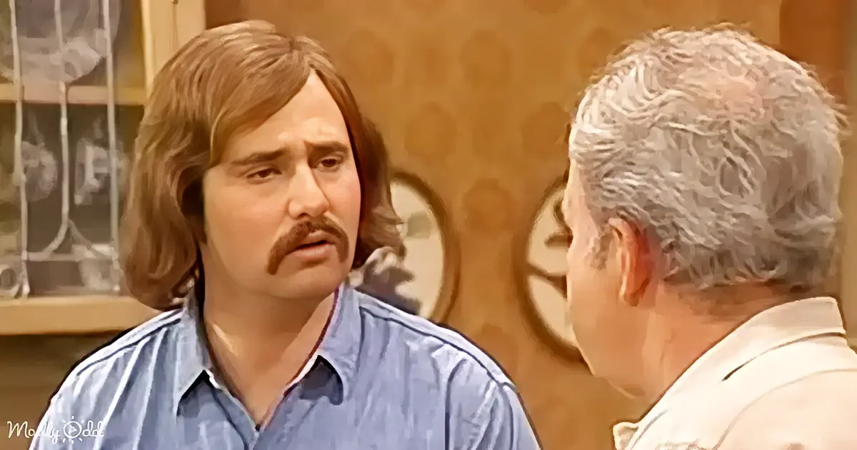 A man with long hair and a mustache in a blue shirt is having a conversation with another man.
