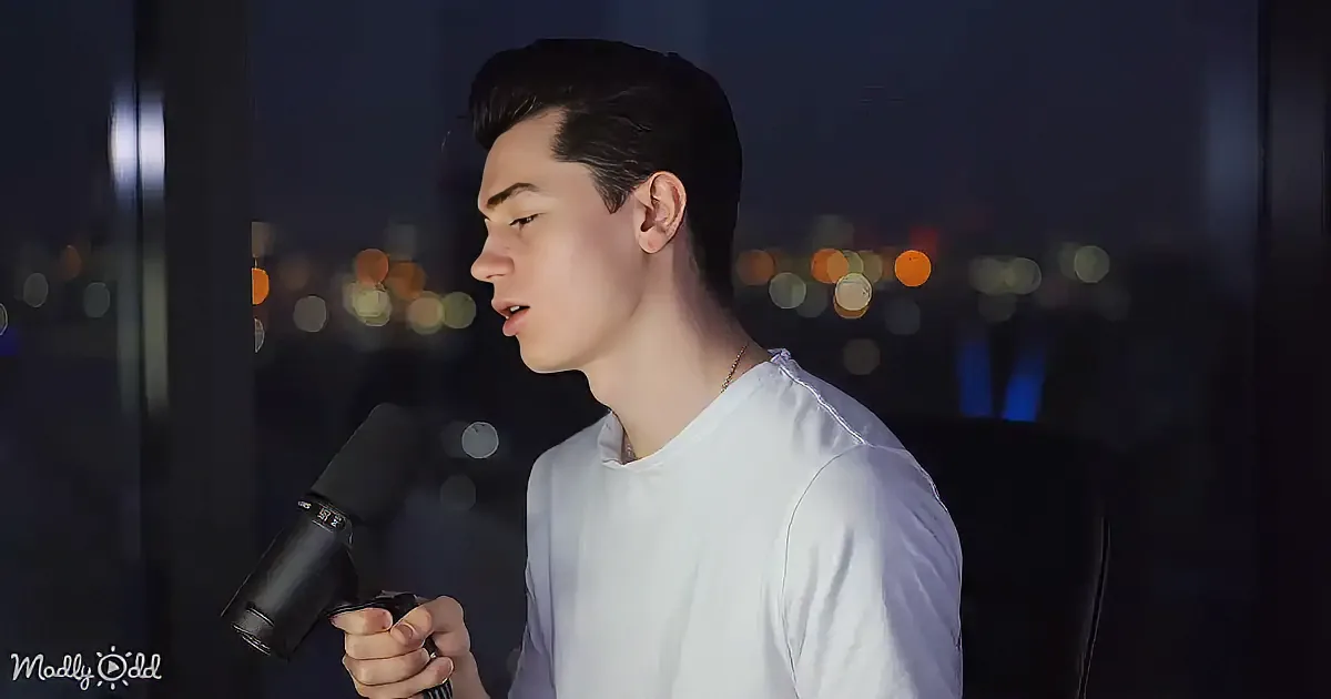 A person in a white shirt holds a microphone, speaking against a cityscape backdrop.
