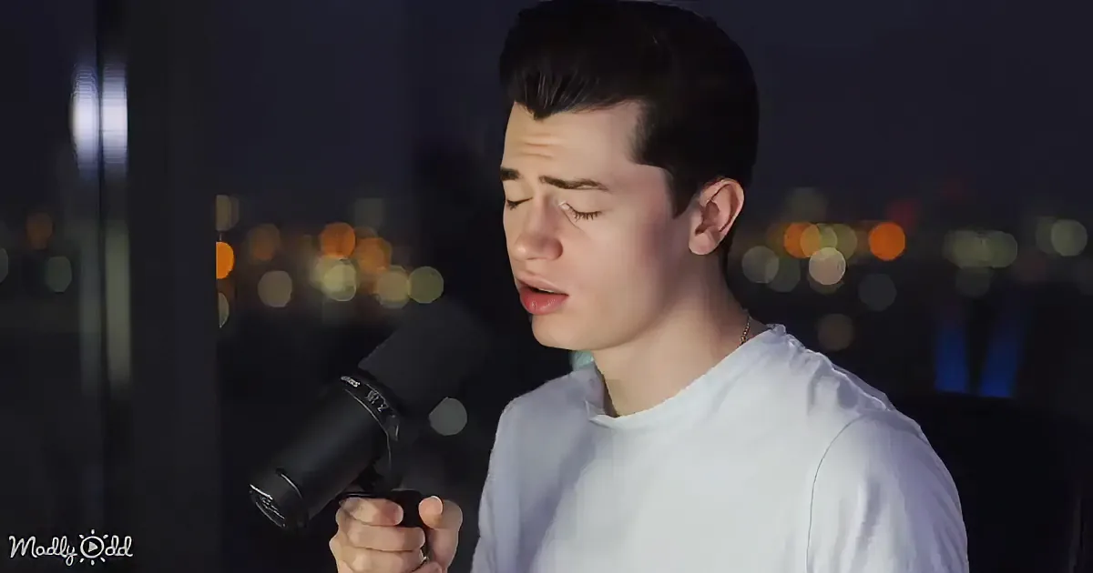 A person in a white shirt sings into a microphone, with a cityscape in the background.