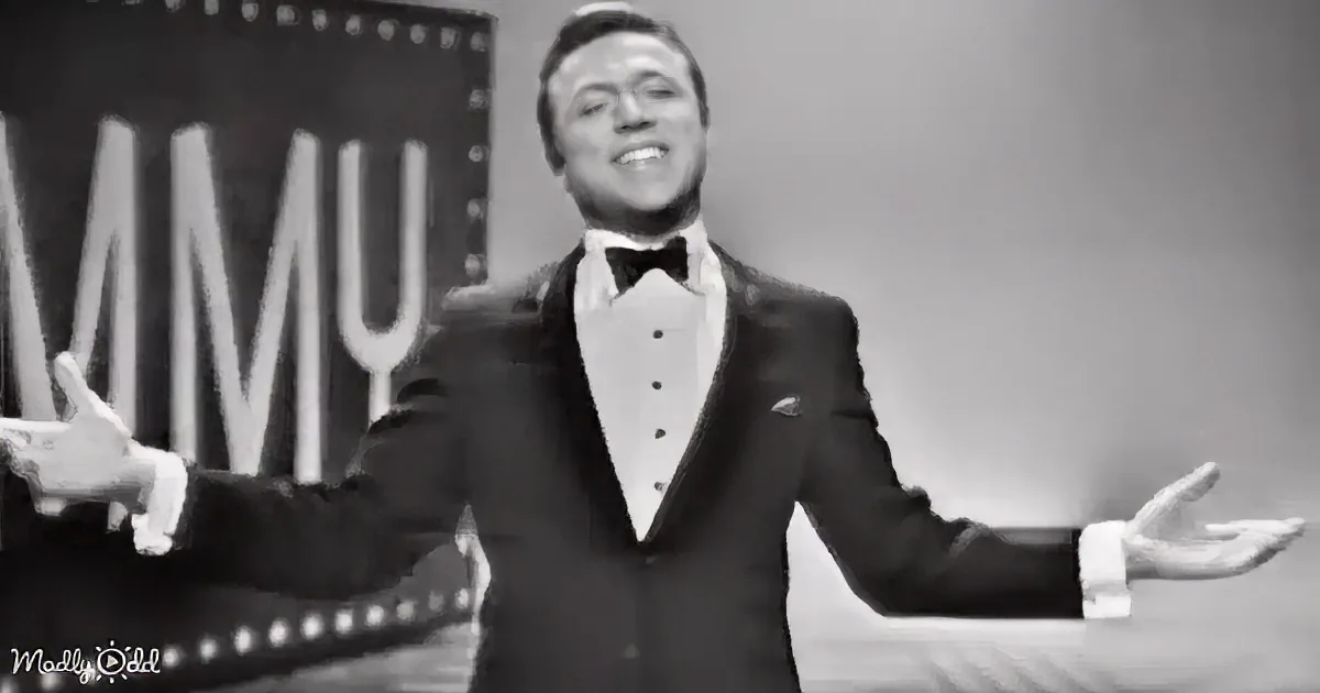 A man in a tuxedo smiles widely, arms outstretched, in front of a "MMY" backdrop.