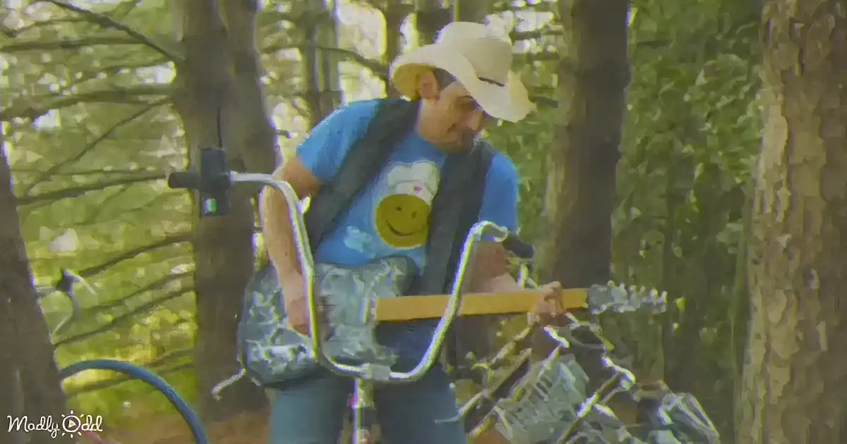 A man in a cowboy hat and blue shirt rides a bicycle in a wooded area.