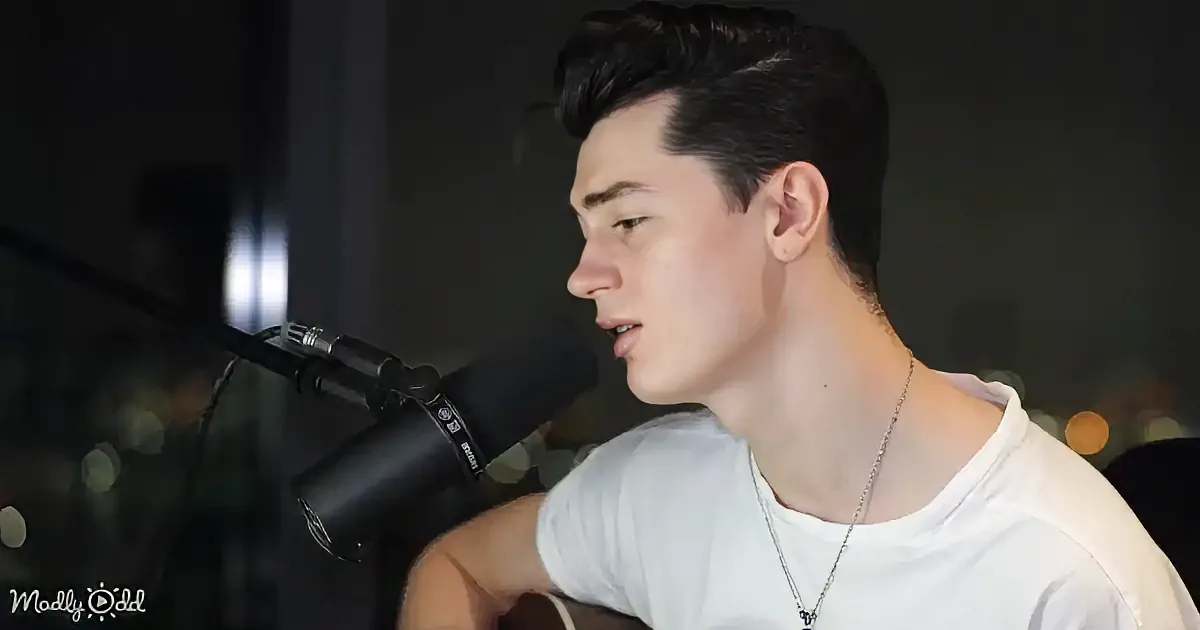 A person in a white shirt sings into a microphone, wearing a necklace, with a blurred background.