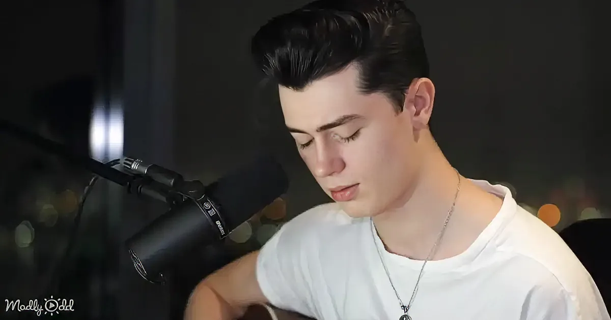 A person in a white shirt sings into a microphone, holding a guitar, with a dark background and blurred lights.