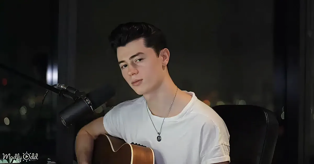 A young man in a white shirt holds an acoustic guitar, sitting in front of a microphone, with a dark background.