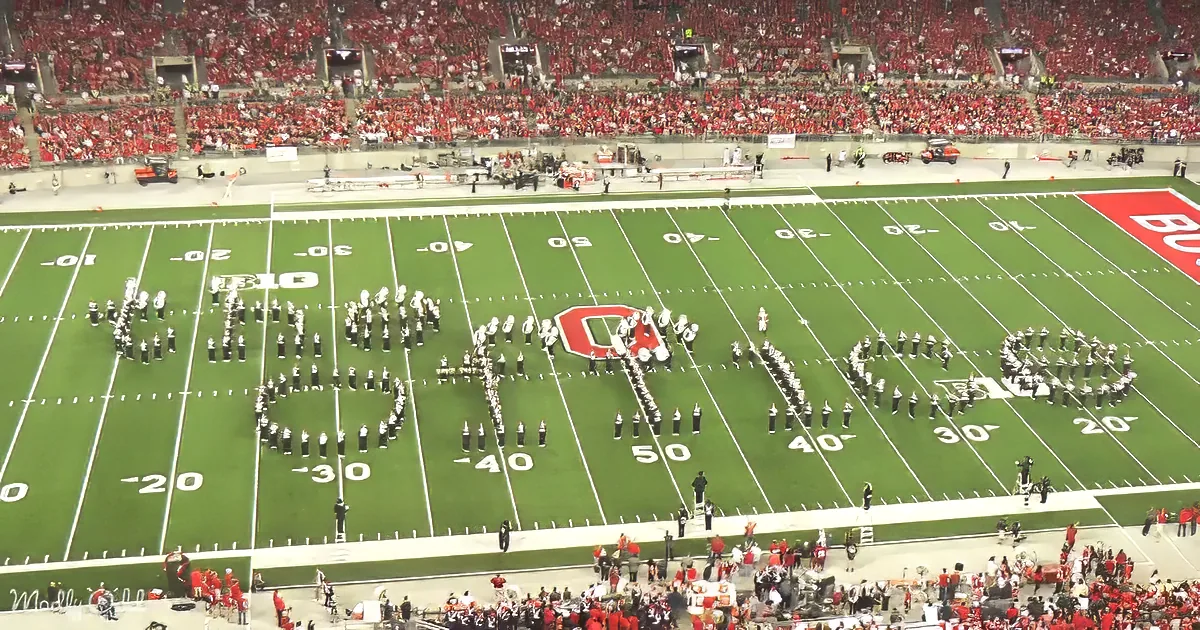 A marching band forms a large "O" on a football field in front of a packed stadium.