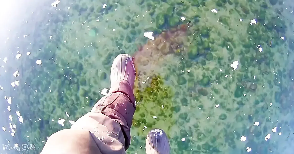 A person wearing boots stands in shallow, clear water with visible rocks and algae beneath.