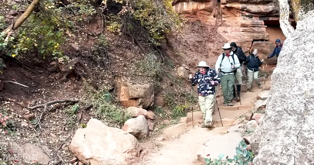 A group of hikers, including one in a floral jacket and hat, ascends a rocky trail surrounded by autumn foliage.
