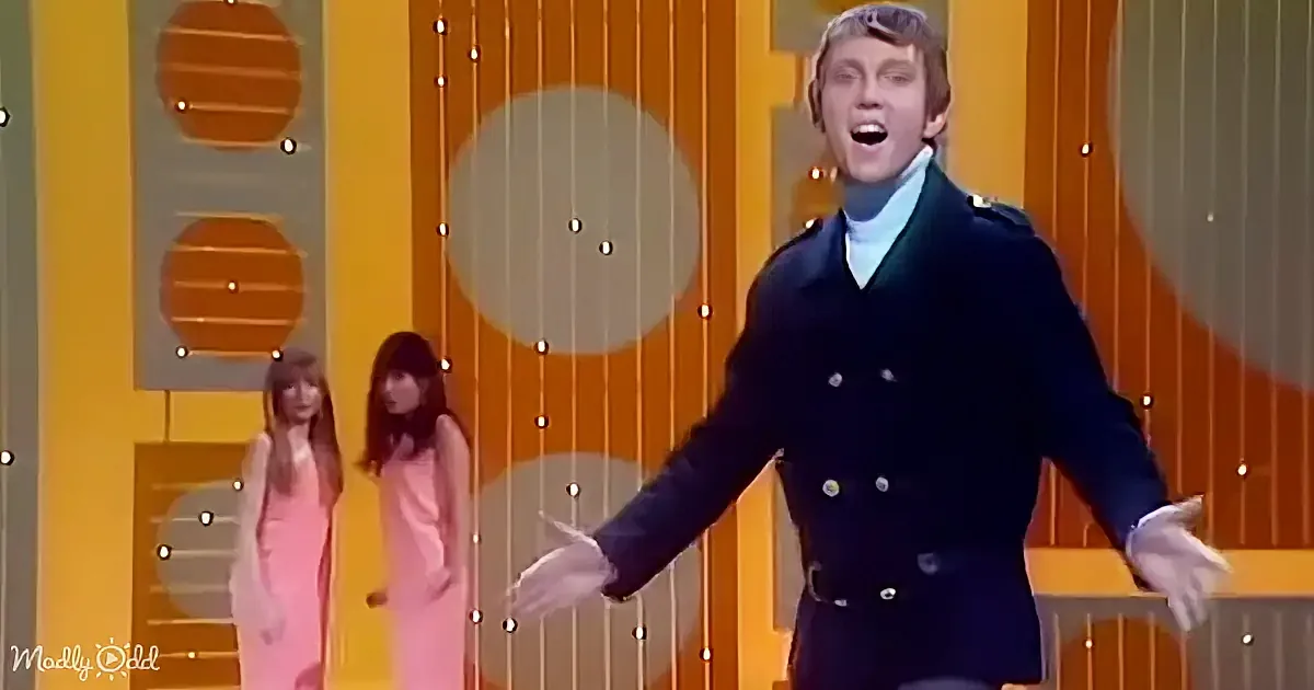 A man in a dark blue double-breasted coat sings on stage with two women in pink dresses behind him.